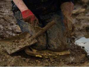 Kneeling in mud clutching archaeological trowel